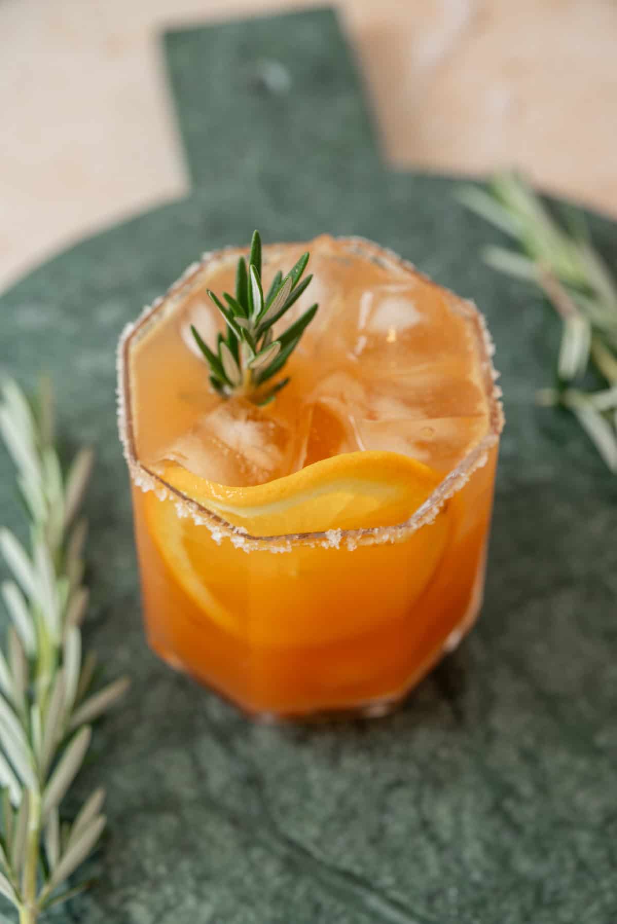 A glass of orange cocktail with ice, garnished with a rosemary sprig and orange slice, sits on a green surface. The glass rim is coated with salt, and additional rosemary sprigs are placed beside it.