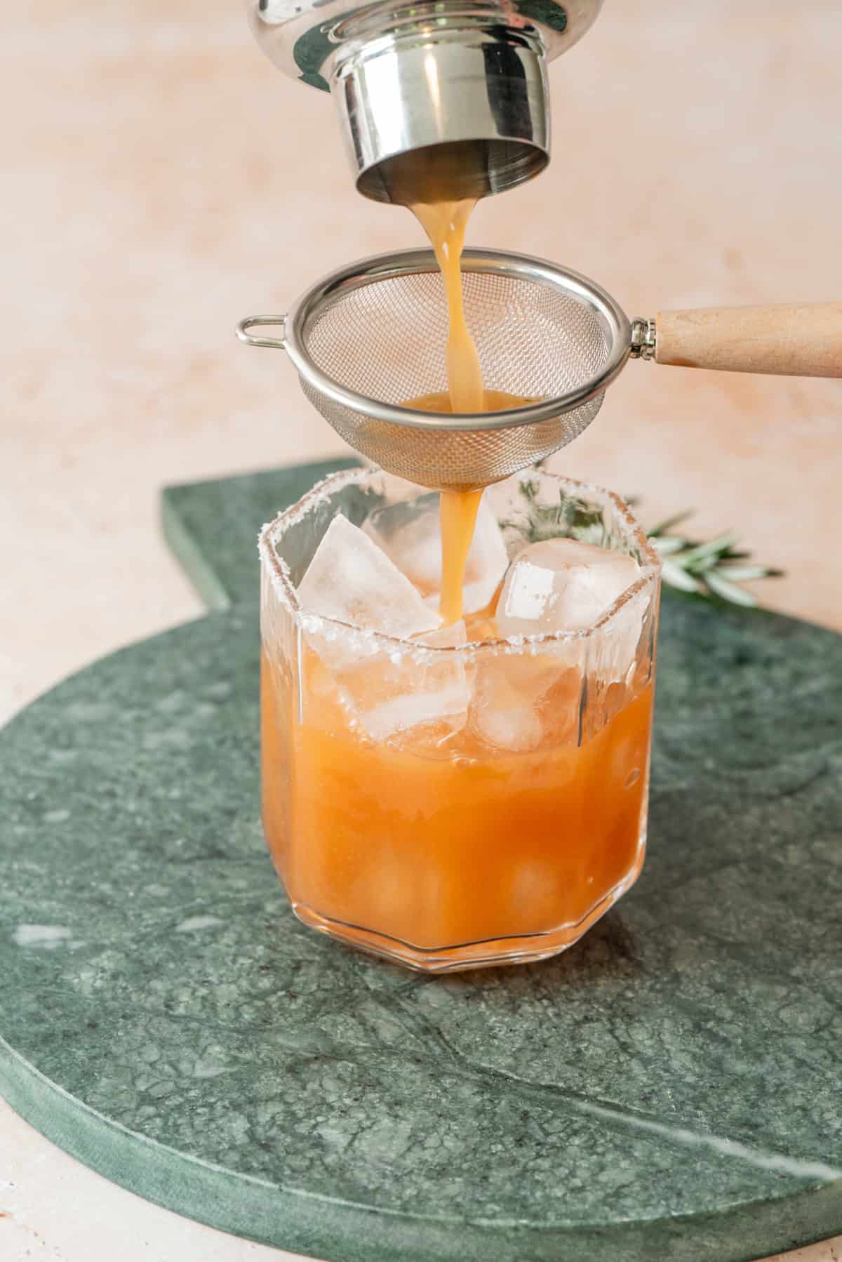 A Cranberry Orange & Rosemary Tonic with ice is being strained into a glass with a salt rim, set on a green stone surface. The drink appears orange in color and a metal strainer is held above the glass.