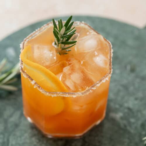 A Cranberry Orange & Rosemary Tonic in a faceted glass with ice cubes, garnished with a rosemary sprig and a citrus peel, and a sugared rim, set on a green marble surface.