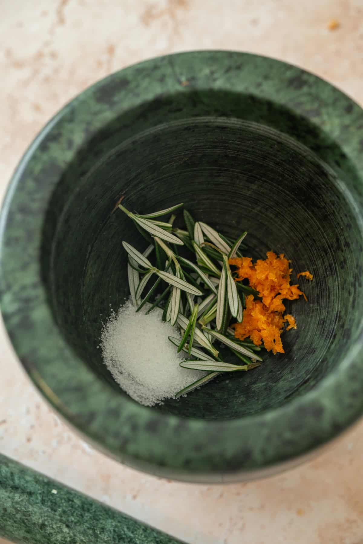 A green marble mortar containing coarse salt, fresh rosemary sprigs, and orange zest, ready to be ground together.
