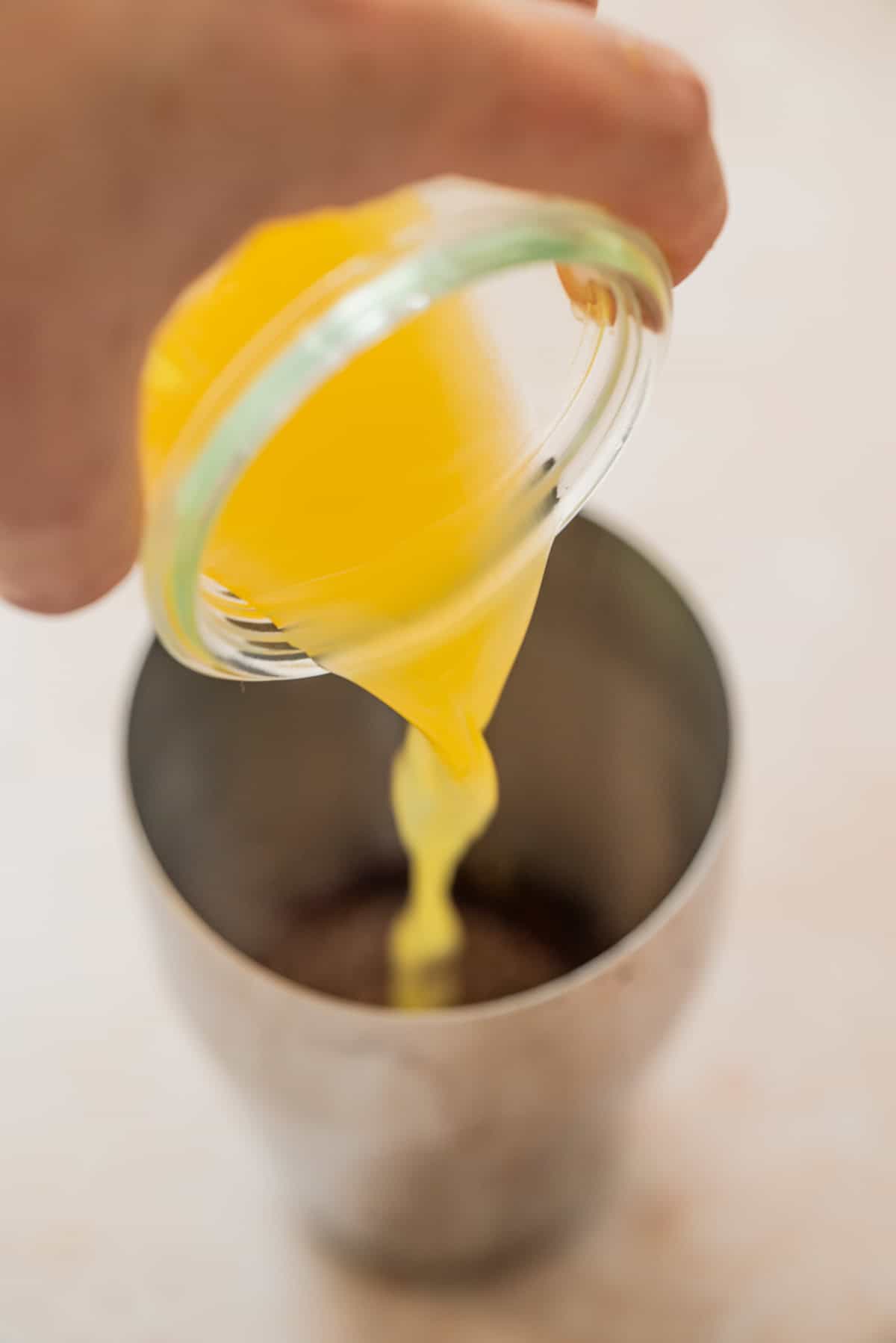 A hand pours a yellow liquid, likely melted butter, from a small glass bowl into a metal mixing cup containing a brown powdery substance.