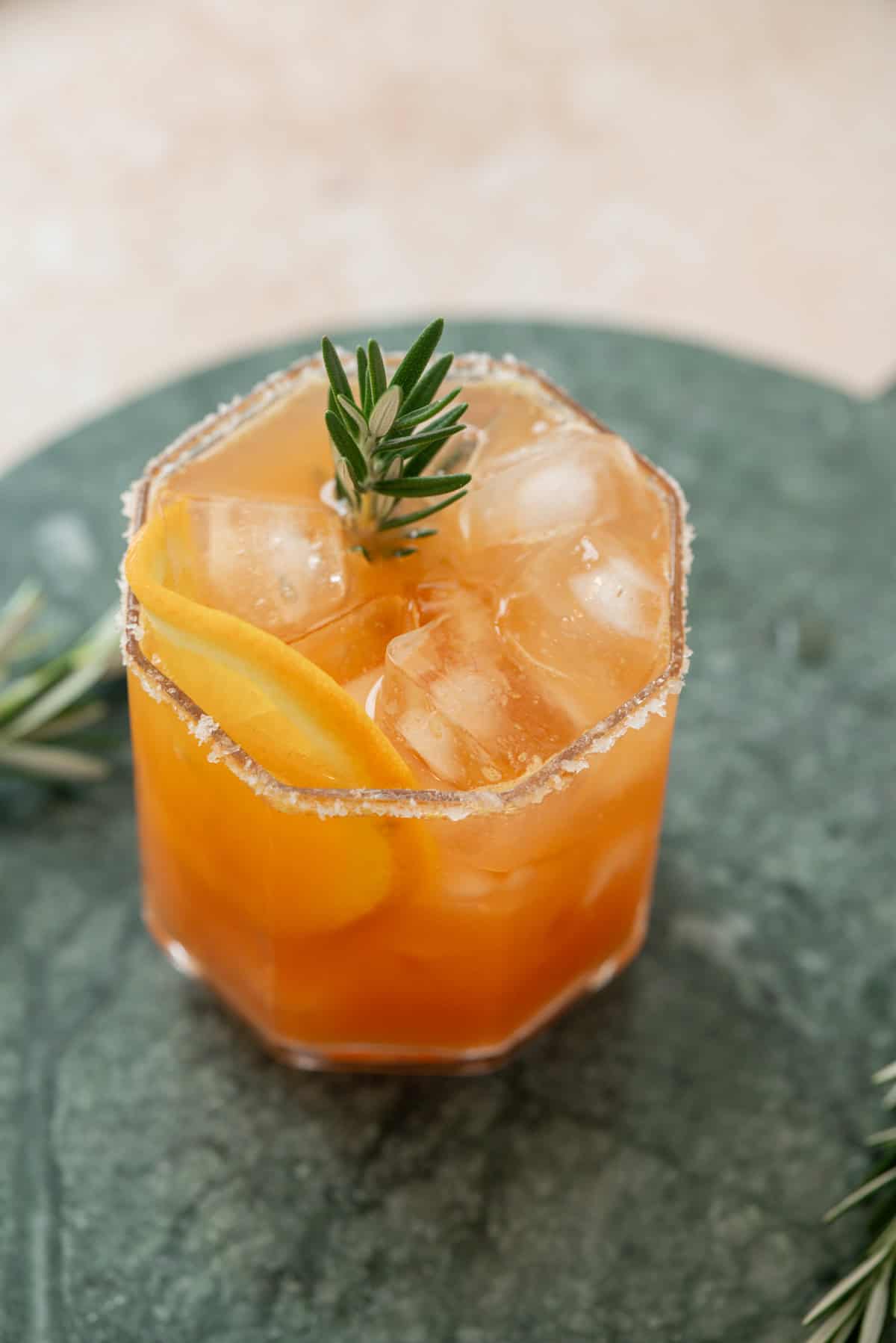 A Cranberry Orange & Rosemary Tonic in a faceted glass with ice cubes, garnished with a rosemary sprig and a citrus peel, and a sugared rim, set on a green marble surface.
