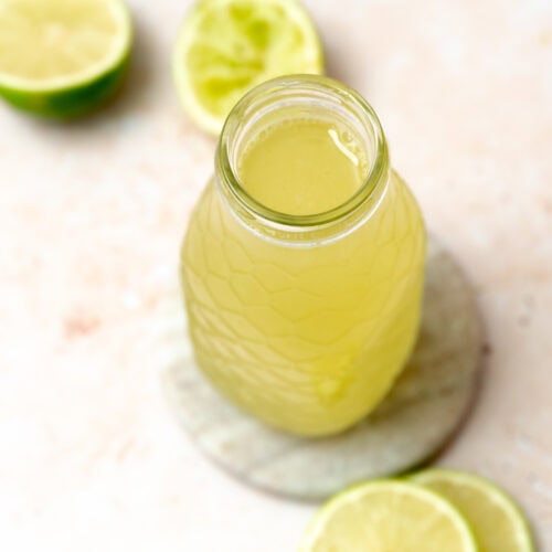 A glass bottle filled with lime cordial sits on a round marble coaster, surrounded by fresh lime slices and lime halves on a light, textured surface.