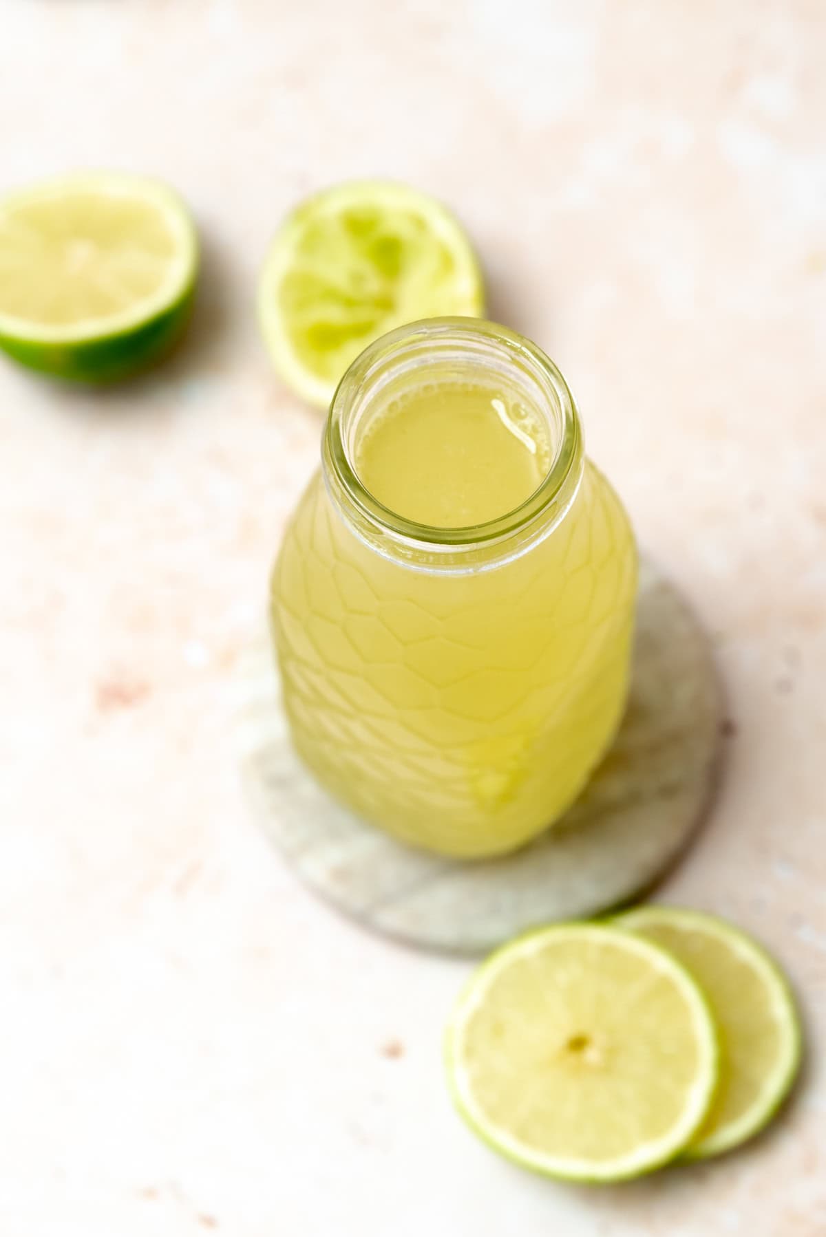 A glass bottle filled with lime cordial sits on a round marble coaster, surrounded by fresh lime slices and lime halves on a light, textured surface.
