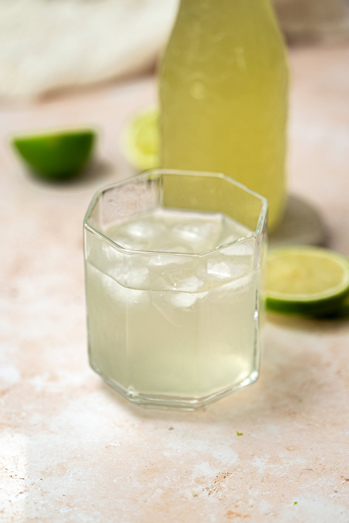 A glass filled with a light-colored, iced beverage sits on a light surface, with lime slices and a bottle in the background.