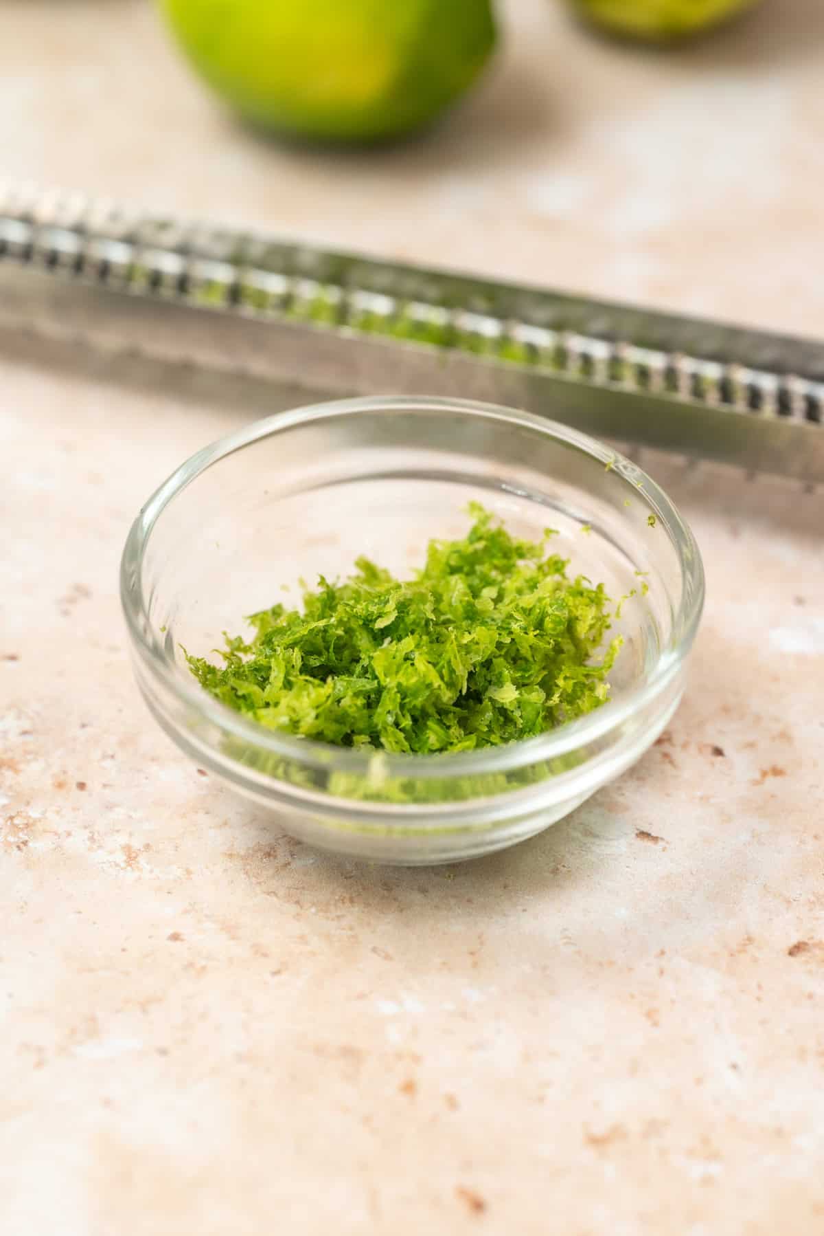 A small glass bowl filled with fresh lime zest sits on a light countertop, with a metal zester and whole limes blurred in the background.