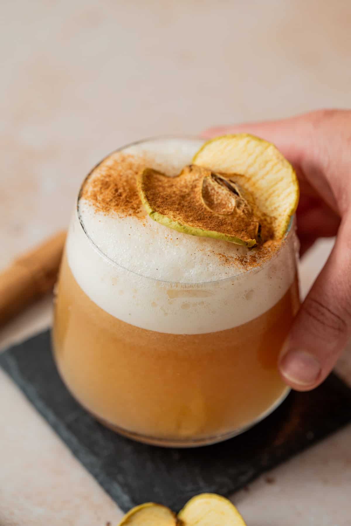 A hand holds a frothy cocktail in a round glass, garnished with a dried apple slice and sprinkled with cinnamon, resting on a dark square coaster.