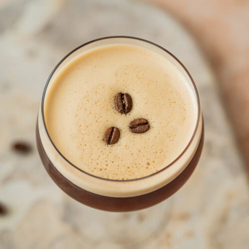 A close-up of a glass filled with a frothy espresso martini, topped with three coffee beans on creamy foam, set on a light, textured surface.