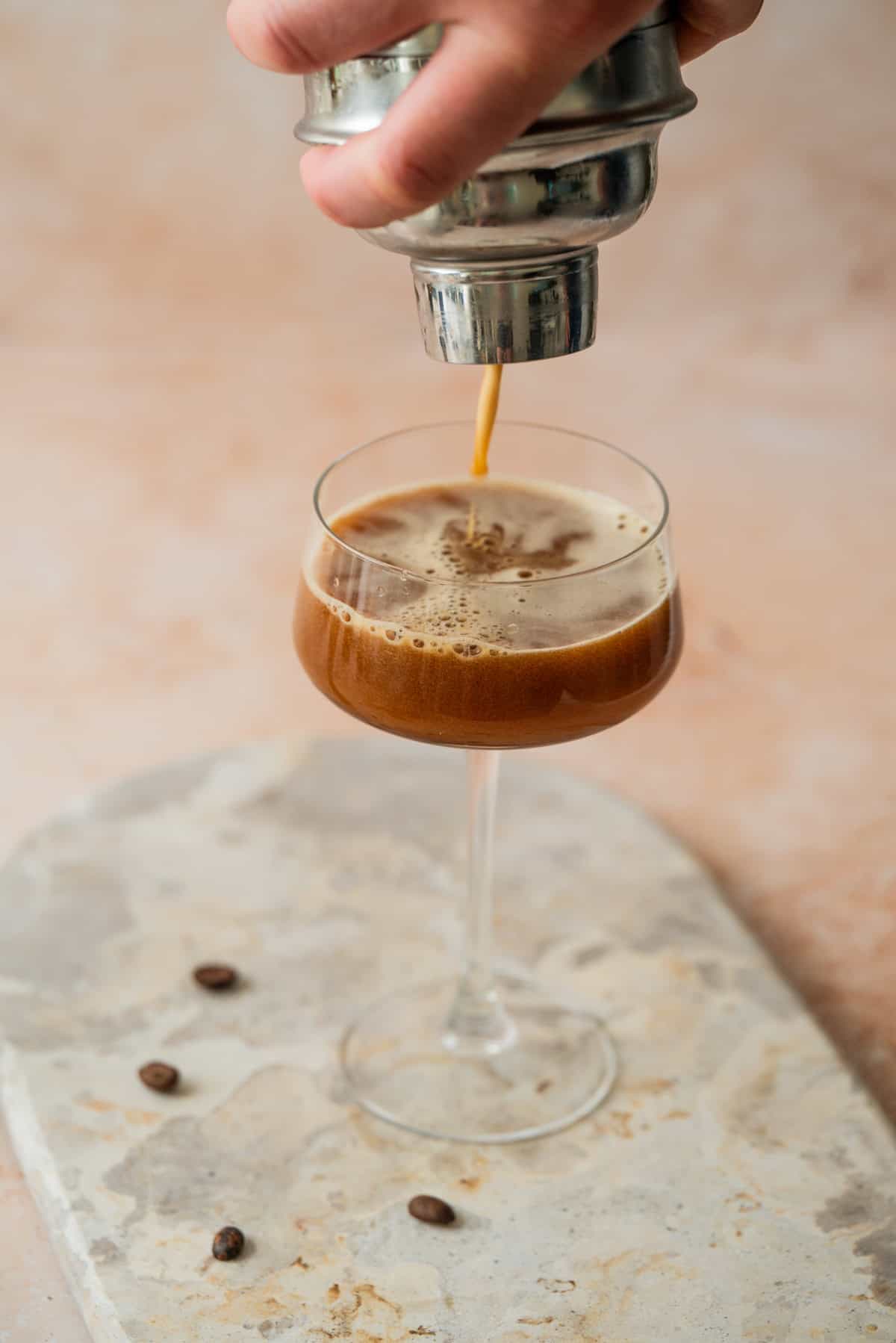 A hand pours a frothy espresso martini from a cocktail shaker into a coupe glass. The glass sits on a light stone surface with coffee beans scattered around.
