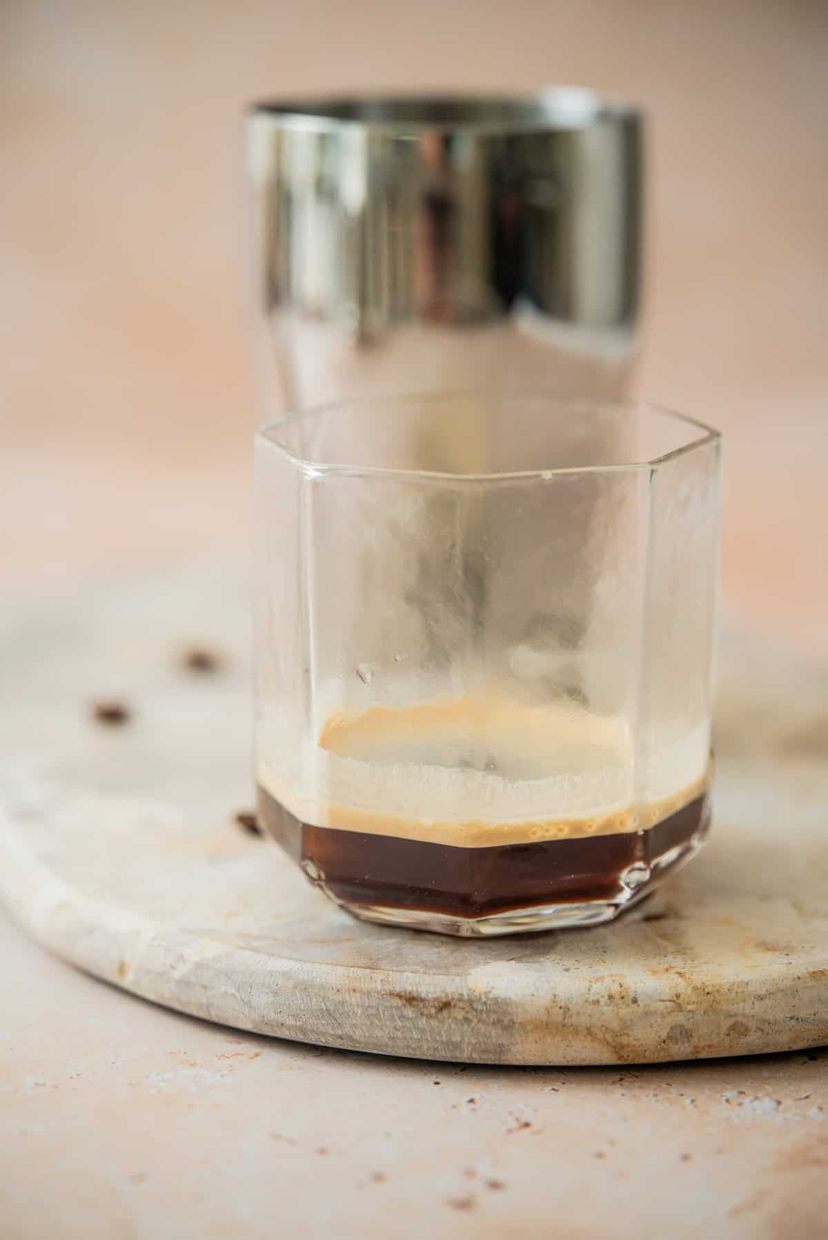 A glass with a small amount of espresso sits on a round wooden board, with a metal shaker and a few coffee beans in the background.
