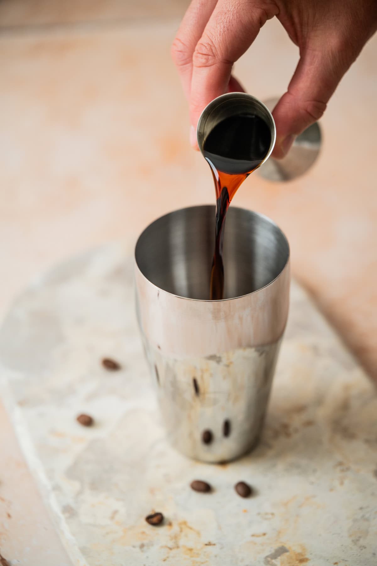 A hand pours a dark liquid from a jigger into a stainless steel cocktail shaker, with a few coffee beans scattered on a light-colored surface nearby.