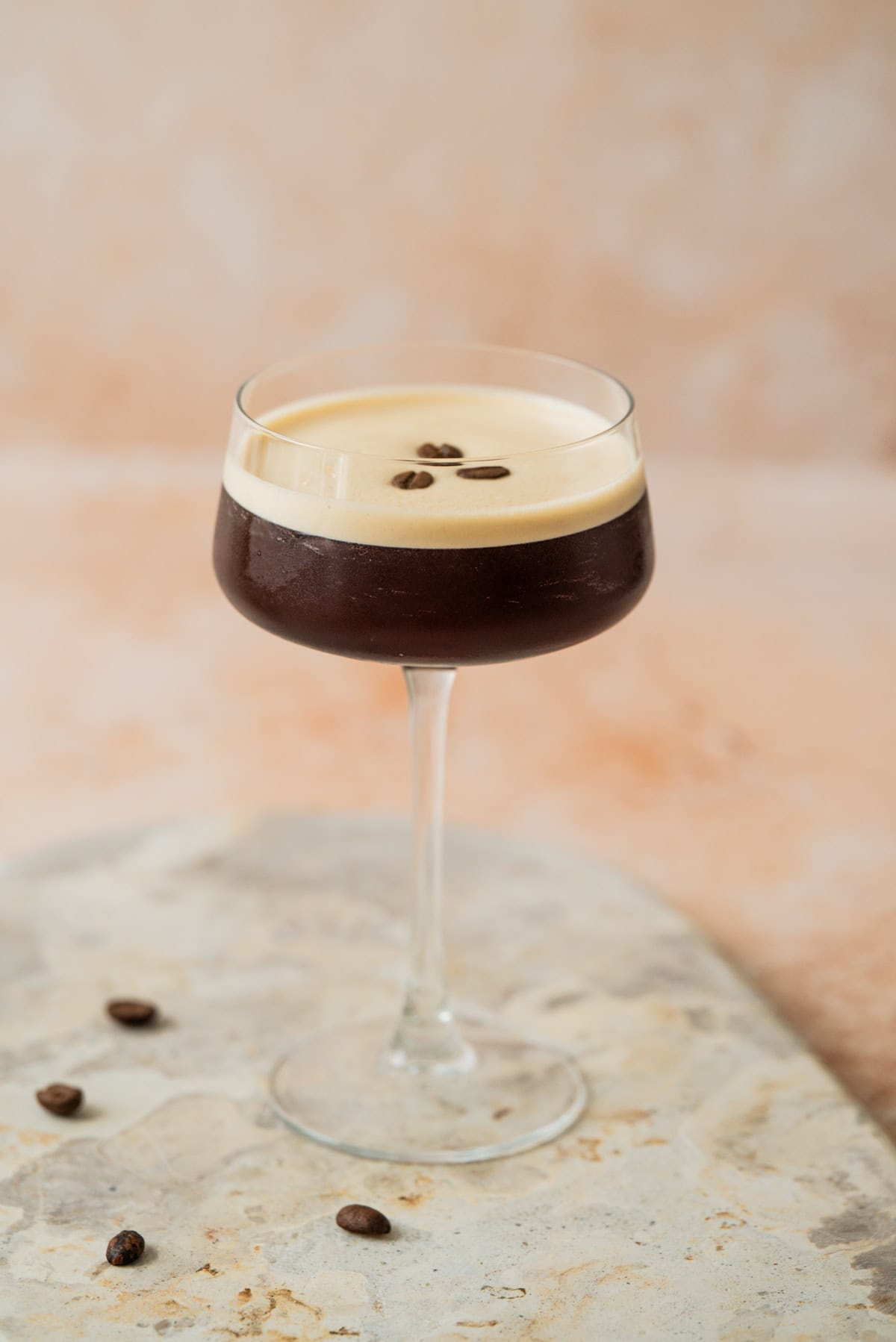 A glass of espresso martini with a creamy foam top, garnished with three coffee beans, is placed on a marbled surface with a few scattered coffee beans around it.