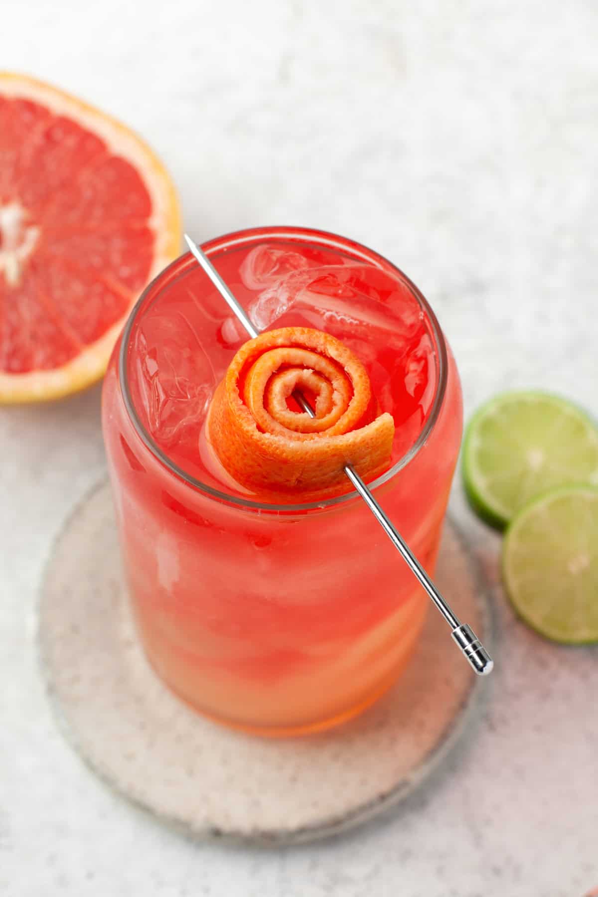 A glass of pink grapefruit cocktail with ice, garnished with a rolled grapefruit peel on a metal pick. Grapefruit and lime slices are placed beside the glass on a light surface.