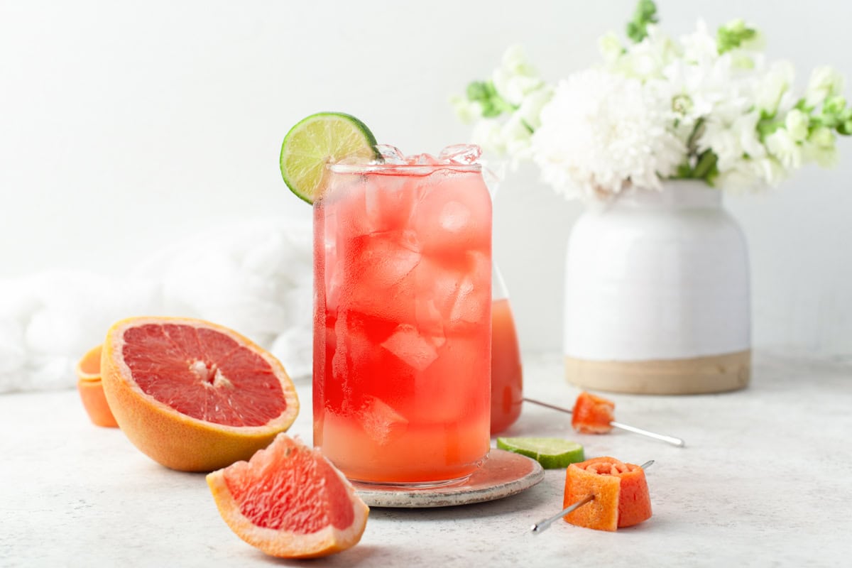 A tall glass of pink grapefruit cocktail with ice and a lime slice sits on a coaster. Fresh grapefruit halves, wedges, and metal skewers are nearby; a white vase with flowers is in the background.