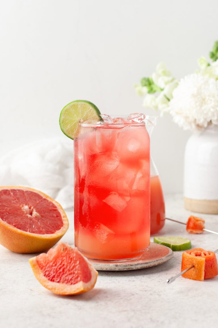 A tall glass of pink grapefruit cocktail with ice is garnished with a lime slice. Fresh grapefruit wedges, lime slices, and a white vase with flowers are in the background on a light surface.