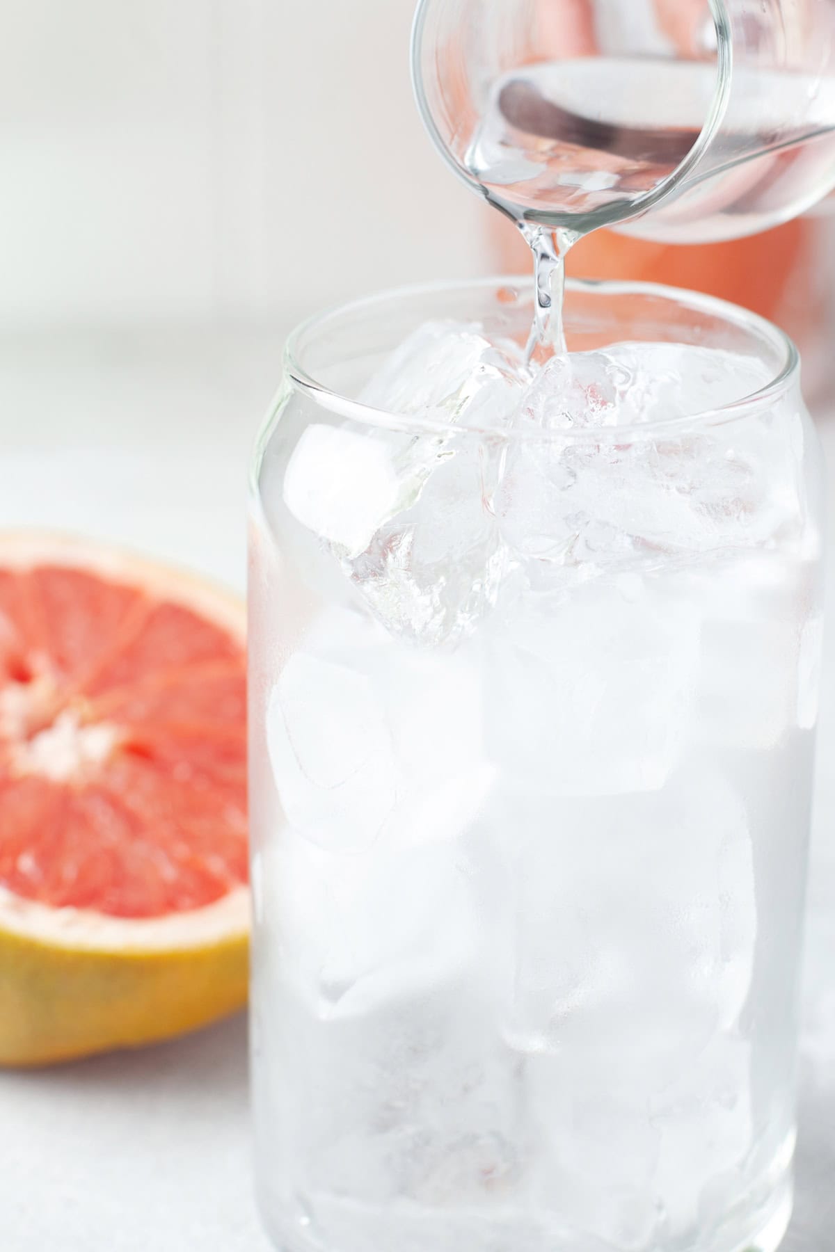A glass filled with ice cubes is being poured with clear liquid from a smaller glass, with a halved grapefruit placed in the background.