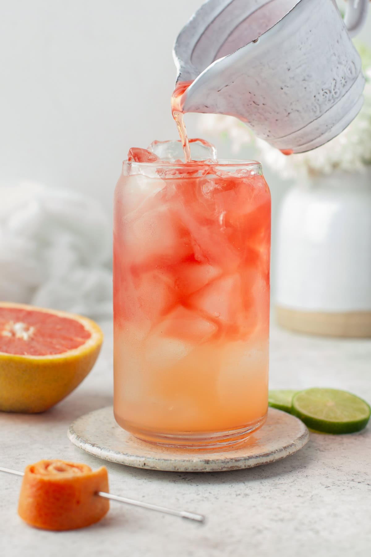 A glass filled with ice and a layered pink and orange drink is being topped off with more liquid from a metal pitcher. A halved grapefruit, a lime slice, and a citrus peel garnish are nearby.