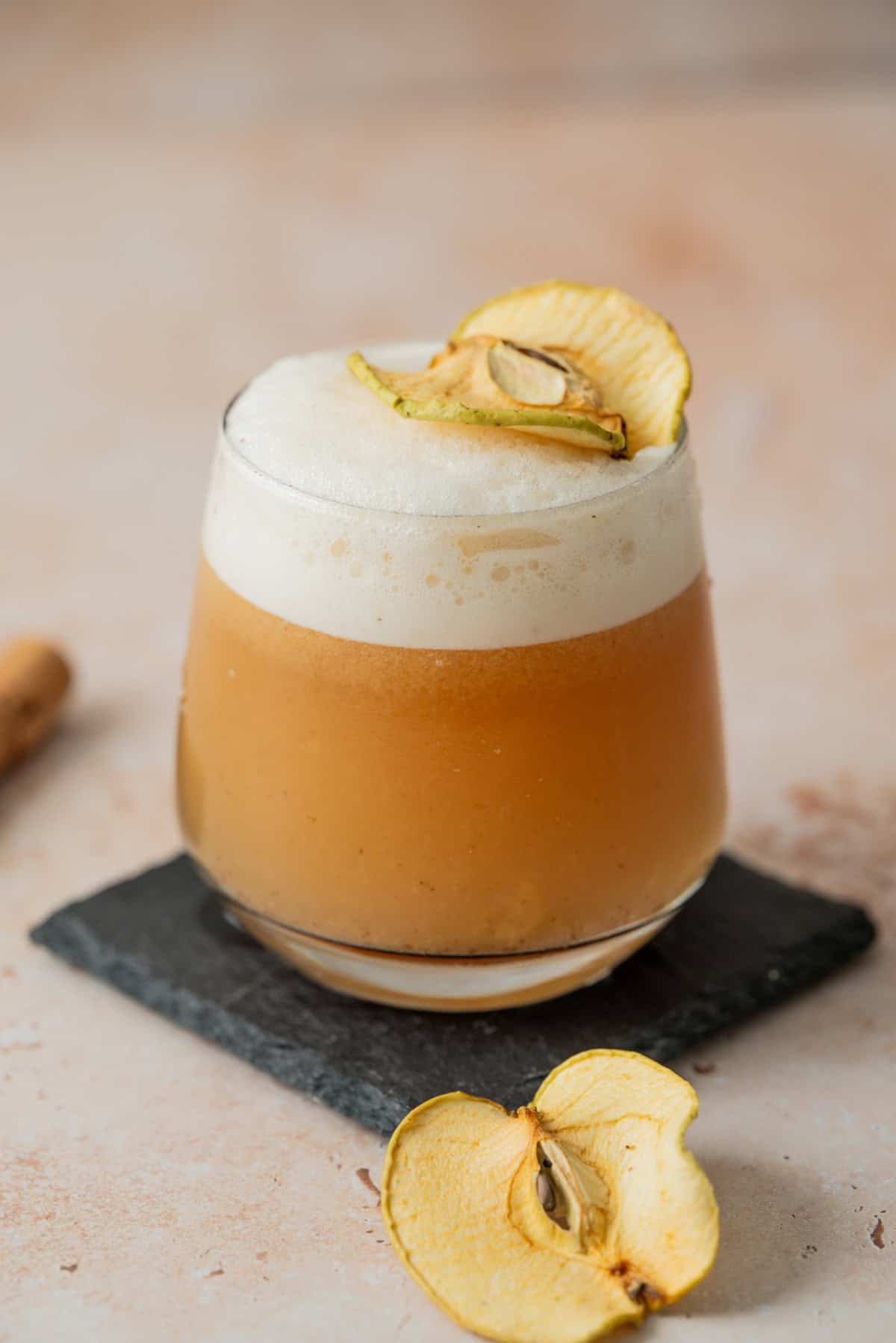 A frothy brown cocktail garnished with dried apple slices, served in a rounded glass on a dark slate coaster, with another dried apple slice on the surface nearby.