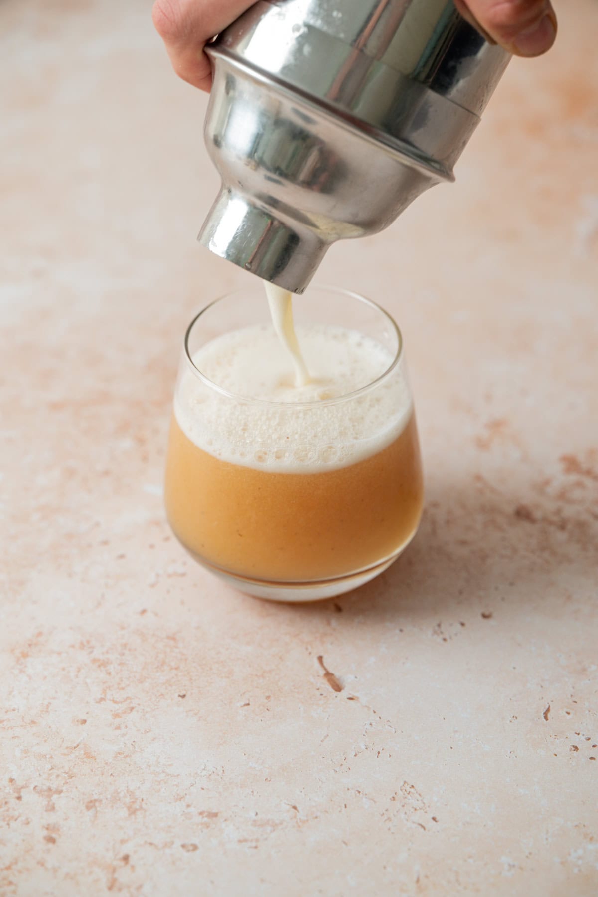 A hand pours a frothy, light brown cocktail from a metal shaker into a rounded glass on a light, textured surface.