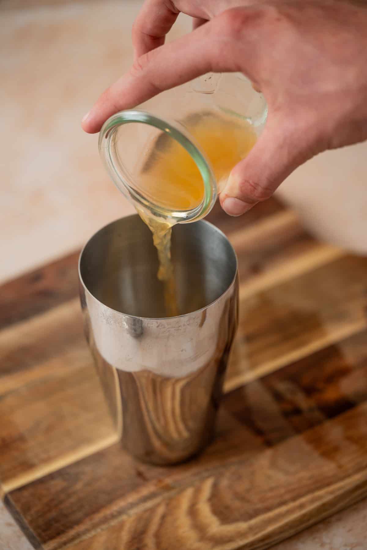 A hand pours orange liquid from a glass jar into a metal cocktail shaker set on a wooden surface.