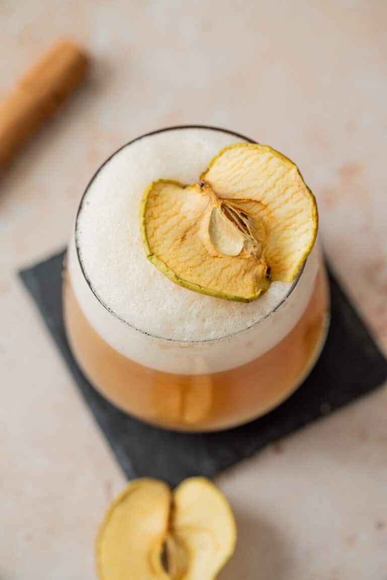 A glass of frothy earl grey sour mocktail garnished with a dried apple slice, placed on a black square coaster with another dried apple slice nearby on a light surface.