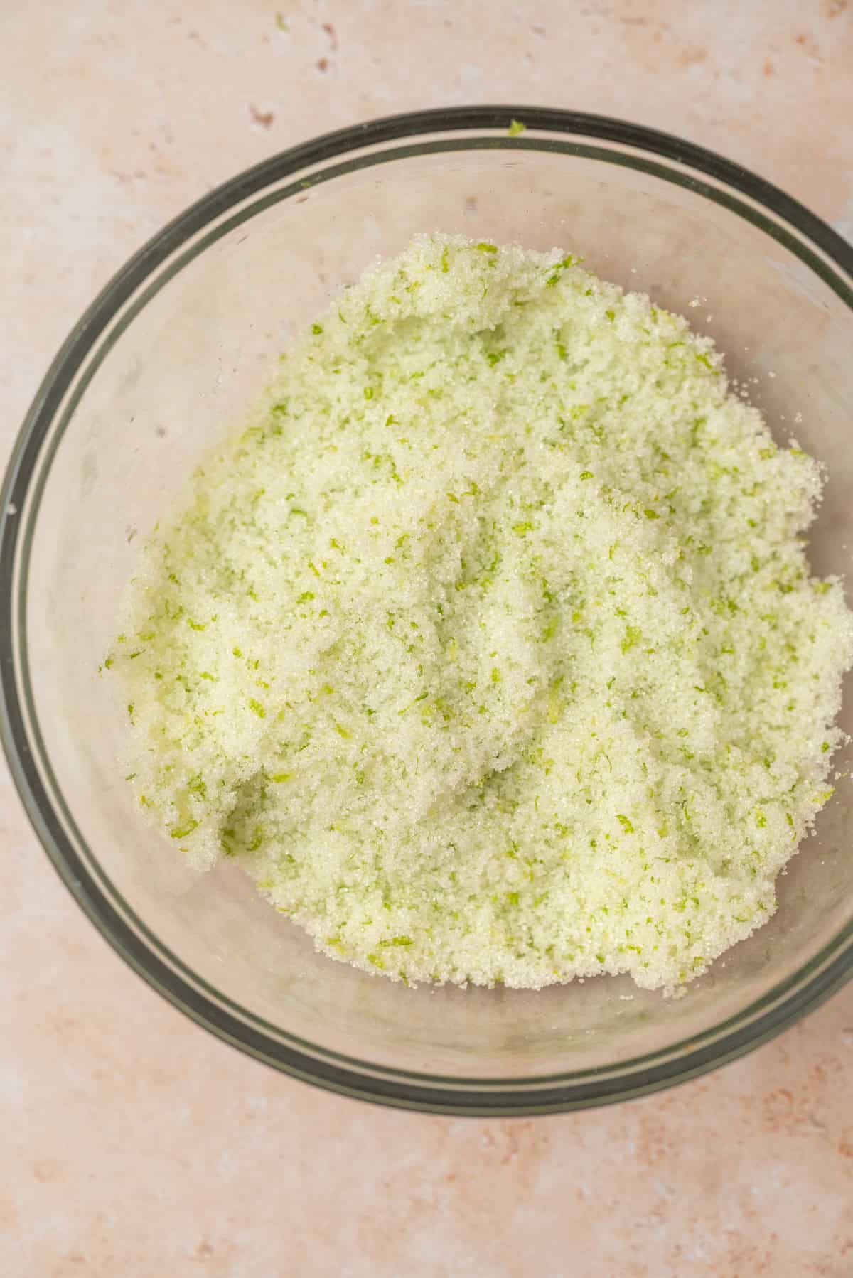 A glass bowl filled with light green, granulated sugar mixed with lime zest, placed on a light beige surface.