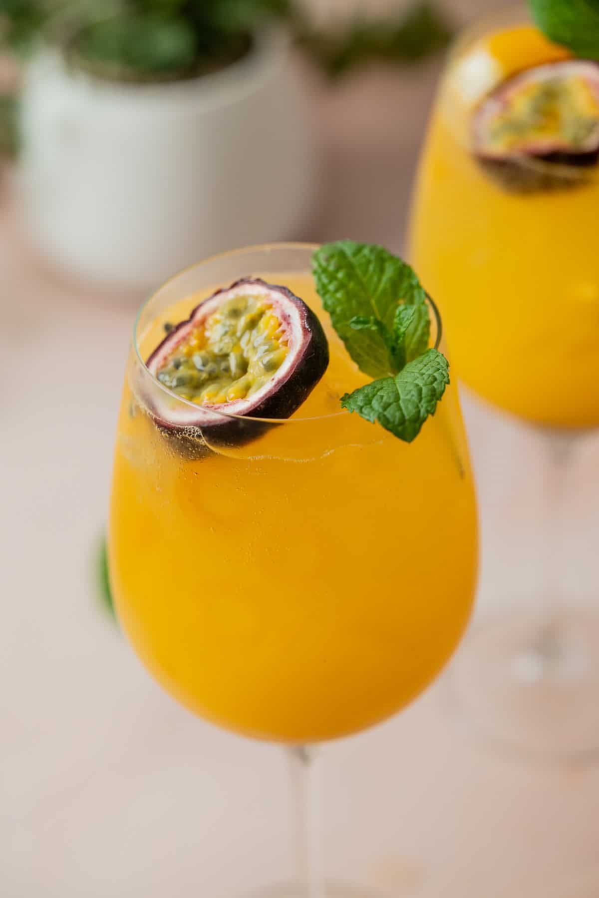 A glass of bright yellow-orange tropical drink, garnished with a halved passion fruit and a sprig of fresh mint. Another similar glass is blurred in the background.