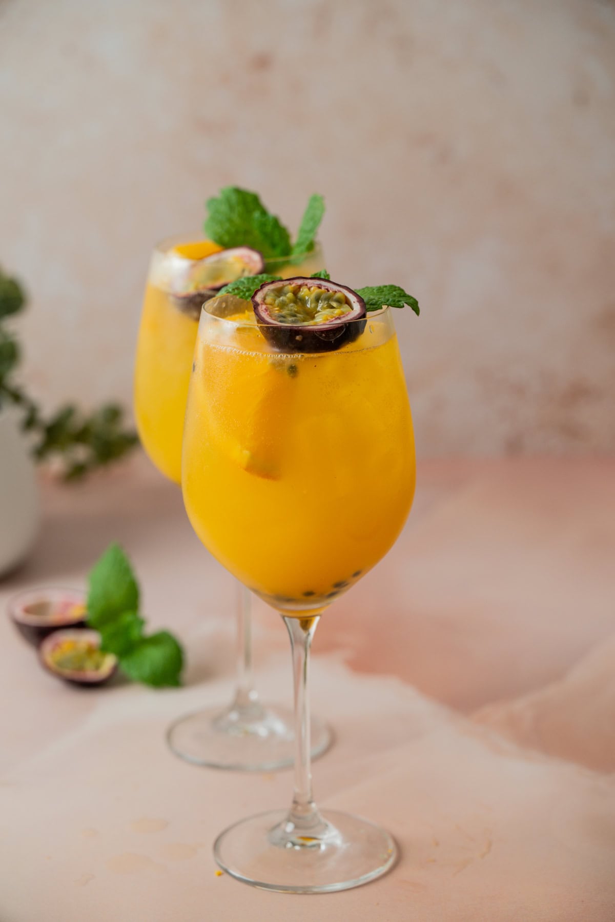 Two elegant wine glasses filled with a bright yellow-orange drink, garnished with halved passion fruit and fresh mint leaves, set on a light pink surface with a soft, neutral background.