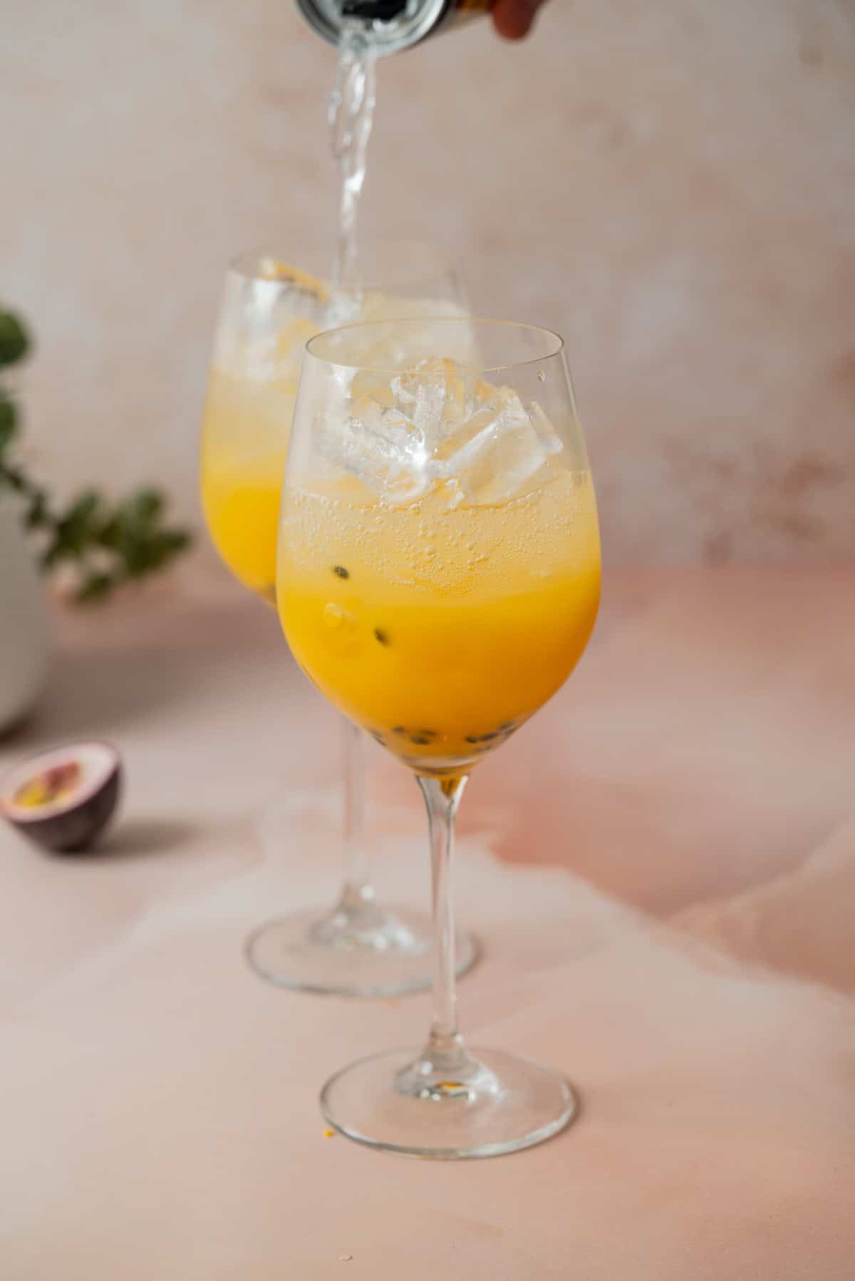 Two wine glasses filled with a yellow passion fruit drink and ice cubes, with a hand pouring liquid into one glass. A halved passion fruit is on the light pink surface in the background.