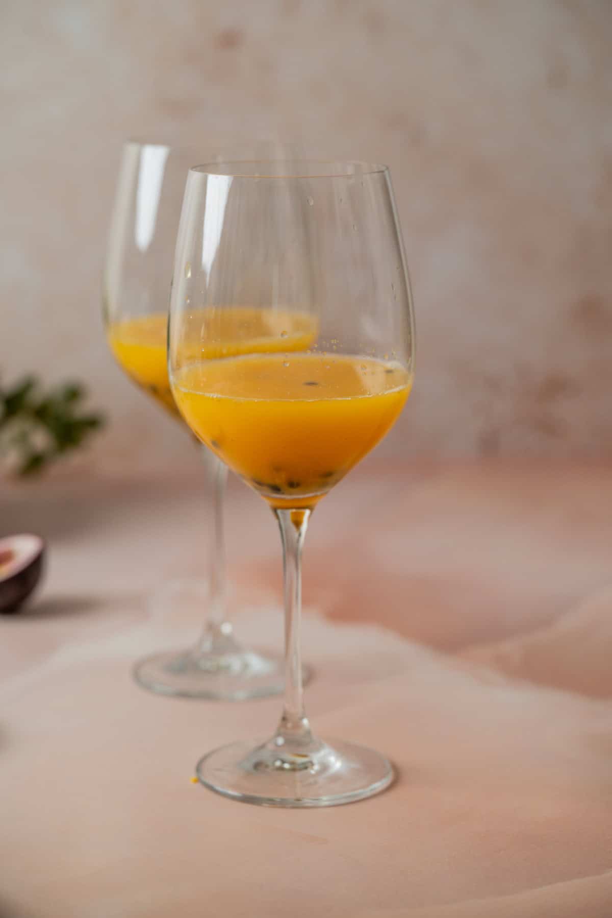Two wine glasses filled with a yellow-orange beverage sit on a pale pink surface, with a blurred background and a partial view of a passion fruit on the left.
