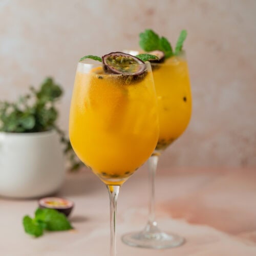 Two glasses of a vibrant yellow passion fruit cocktail with ice, garnished with mint leaves and a halved passion fruit. A blurred plant and extra mint leaves appear in the background on a light pink surface.