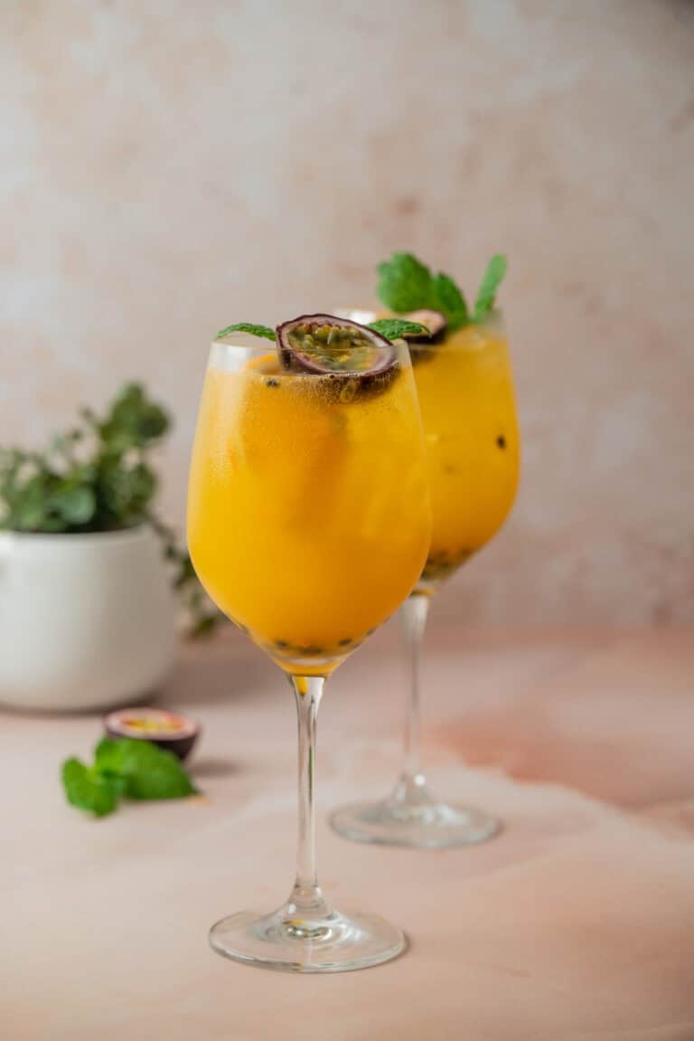 Two glasses of a vibrant yellow passion fruit cocktail with ice, garnished with mint leaves and a halved passion fruit. A blurred plant and extra mint leaves appear in the background on a light pink surface.