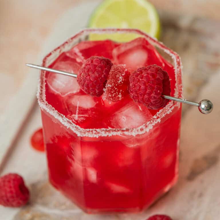 A vibrant Raspberry Cranberry Mocktail with ice cubes in a glass rimmed with sugar, garnished with fresh raspberries on a metal pick, and a lime slice in the background.