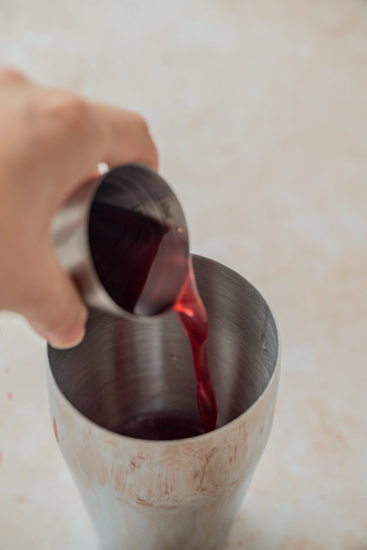 A hand pours a deep red liquid from a metal jigger into a stainless steel cocktail shaker on a light-colored surface.