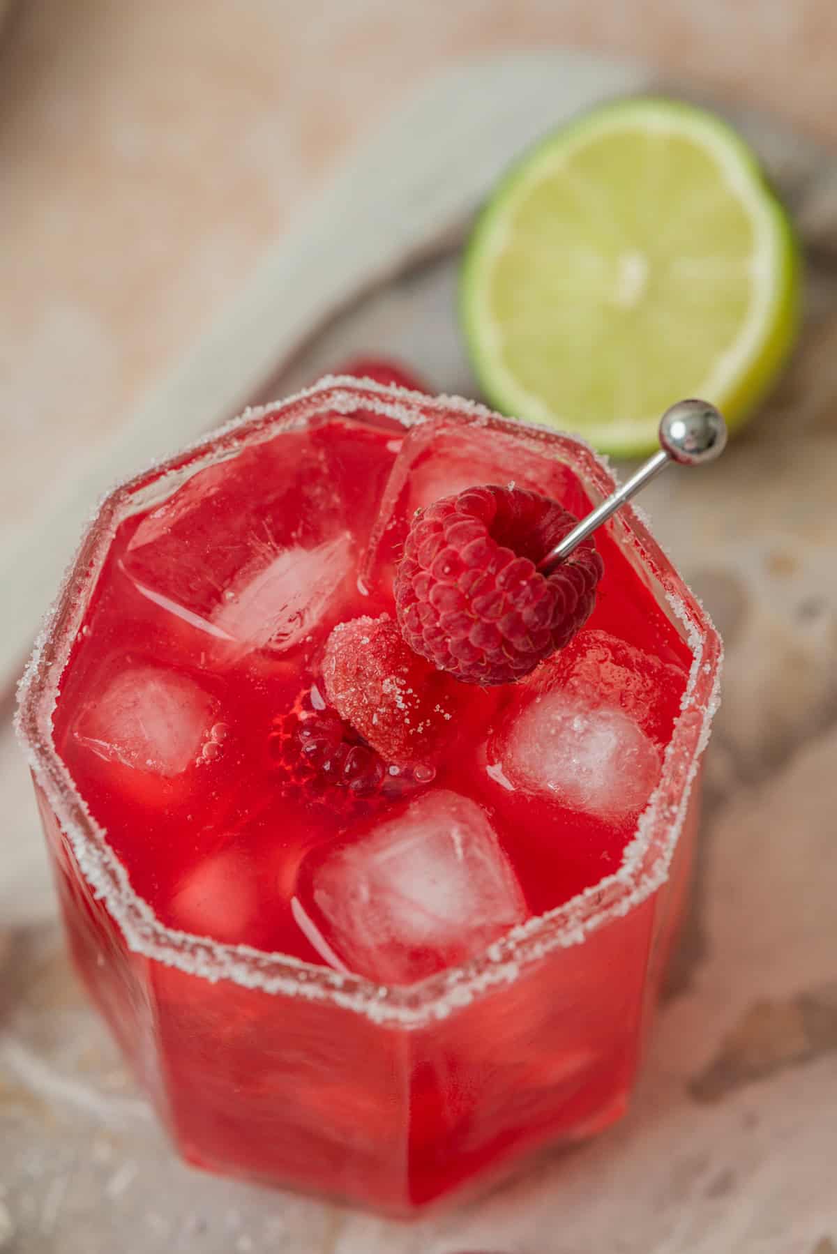 A red cocktail with ice cubes in a glass rimmed with sugar, garnished with fresh raspberries on a cocktail pick. A halved lime is in the background on a light-colored surface.