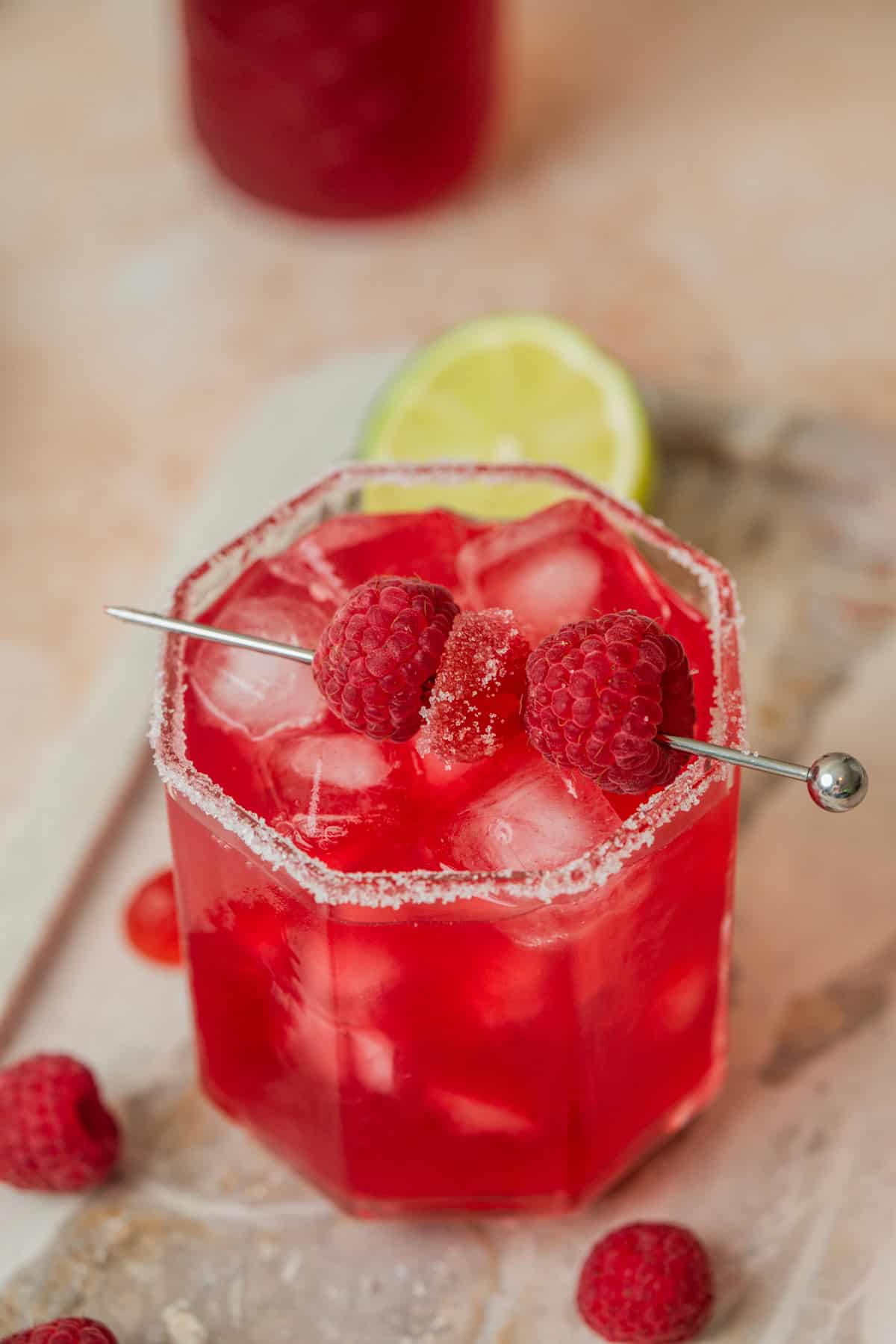 A vibrant red raspberry cranberry mocktail with ice in a salt-rimmed glass, garnished with two raspberries and a sugar cube on a metal pick. Fresh raspberries and a lime slice are nearby on a light marble surface.
