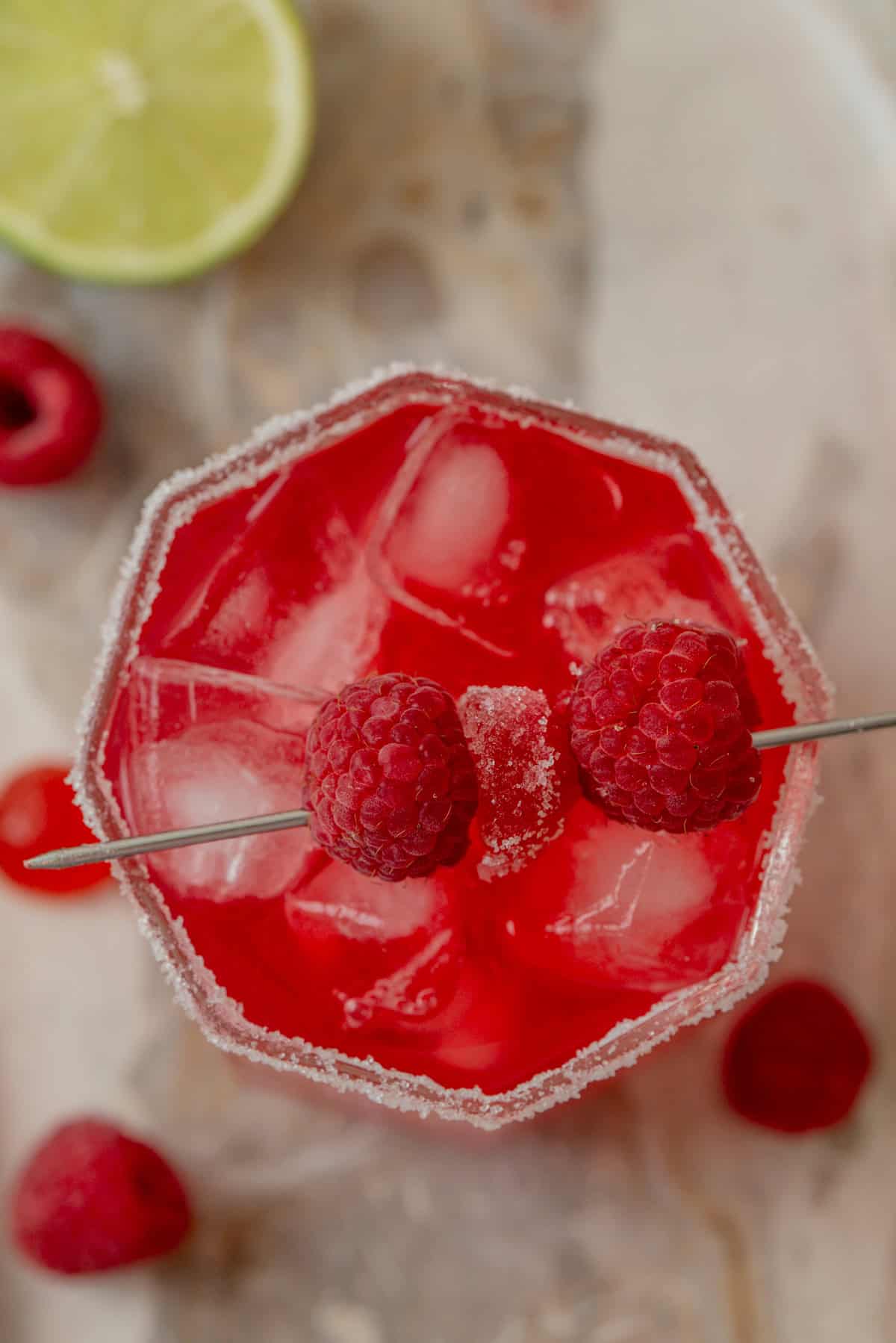 A top-down view of a raspberry cranberry mocktail with ice, garnished with two raspberries on a skewer, in a sugar-rimmed glass. Fresh raspberries and a slice of lime are nearby on a marble surface.