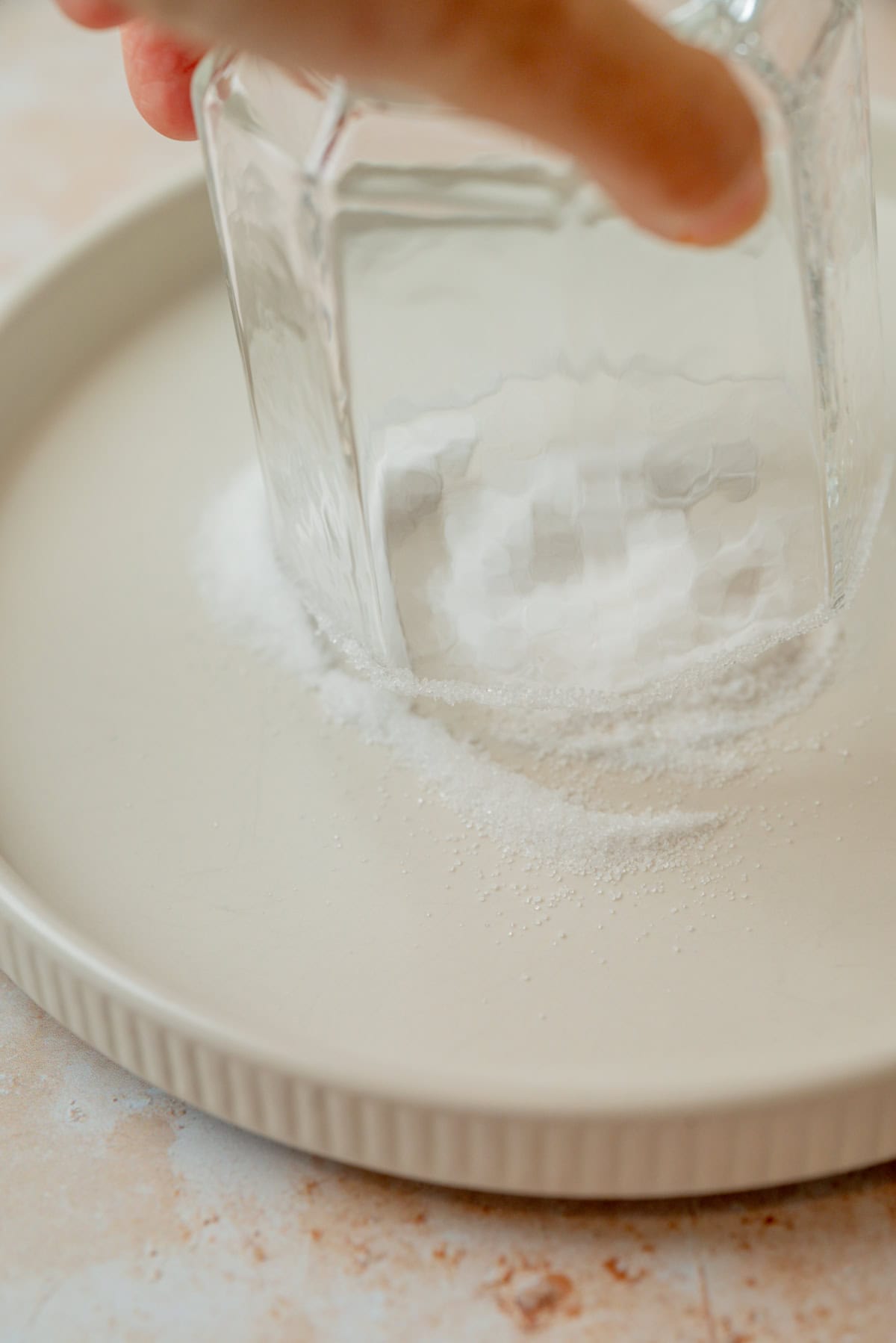 A hand presses a glass upside down onto a shallow plate sprinkled with salt, likely preparing the glass for a salted rim.