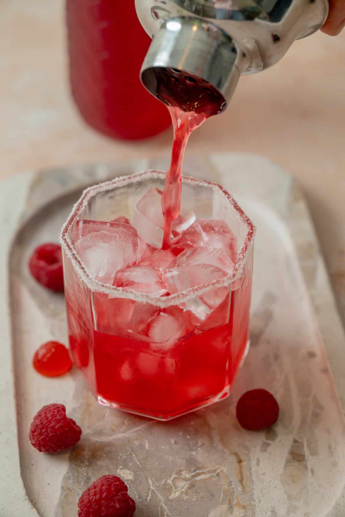 A vibrant raspberry cranberry mocktail is being poured from a shaker into an octagonal glass filled with ice, garnished with a sugar rim, and surrounded by fresh raspberries on a marble tray.