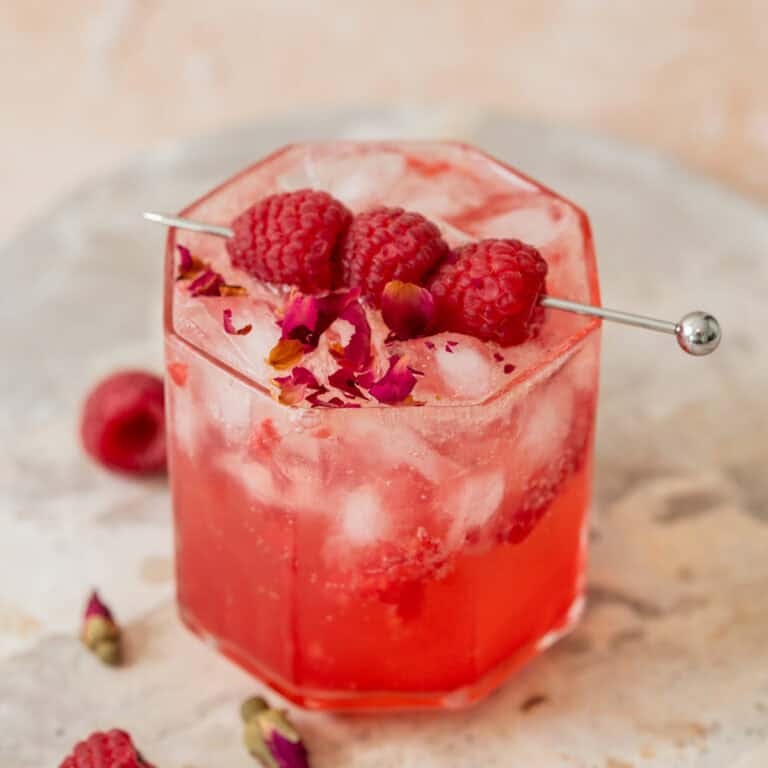 A pink iced raspberry mocktail in a faceted glass, garnished with three raspberries on a metal pick and sprinkled with rose petals, sits on a light stone surface with a few raspberries and rose buds nearby.