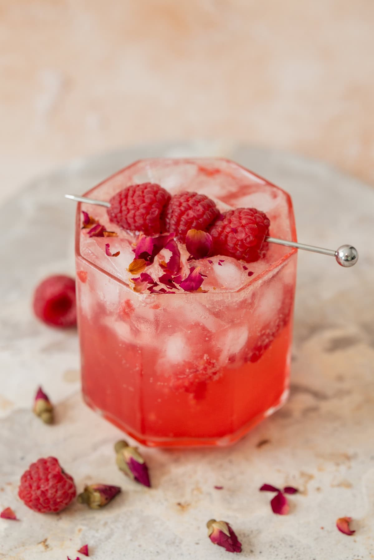 A pink raspberry rose tonic mocktail with ice, garnished with fresh raspberries and rose petals, served in a faceted glass with a metal pick, sits on a light surface with scattered raspberries and rose petals around it.