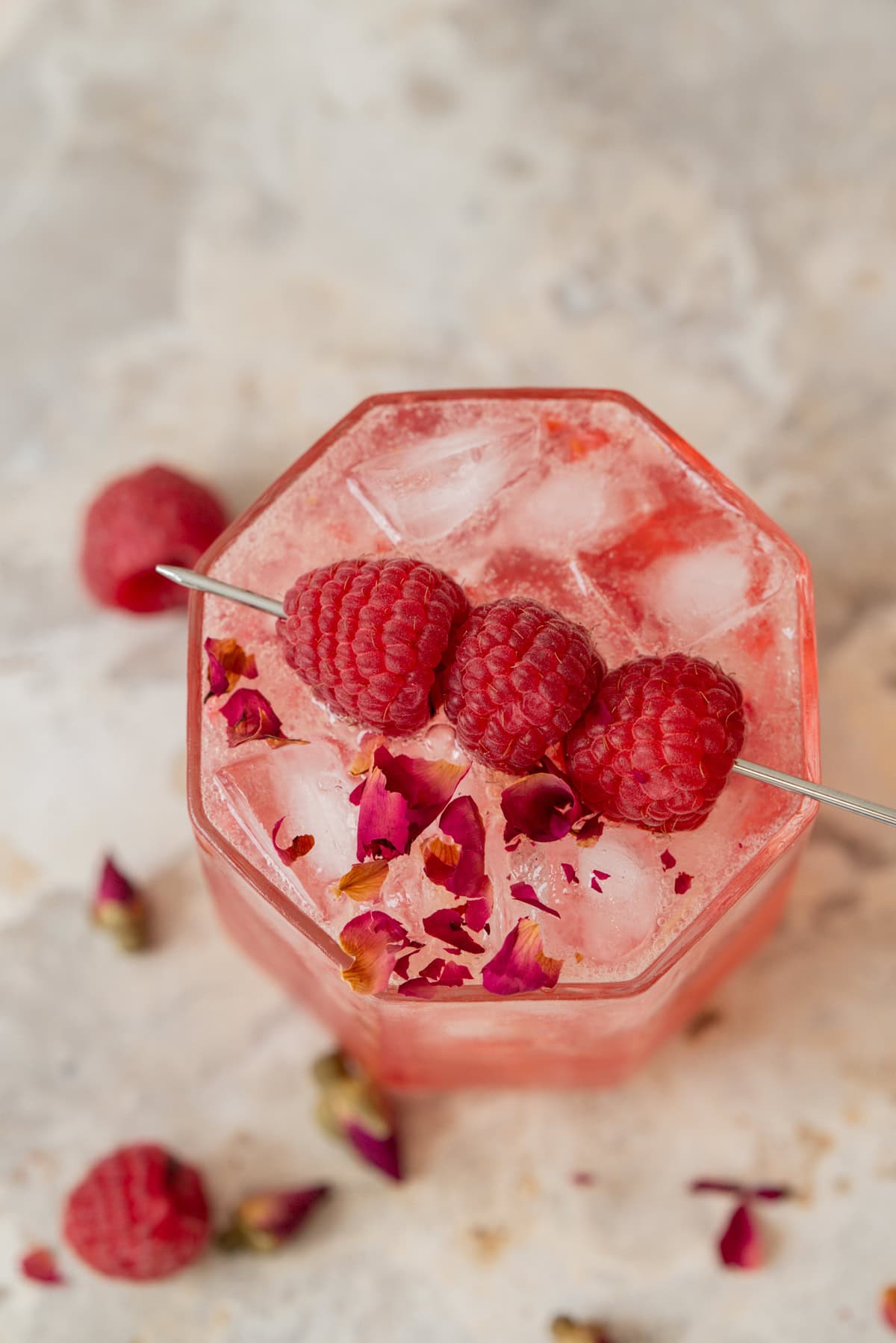 A glass filled with ice and a pink drink, garnished with three fresh raspberries on a skewer and sprinkled with rose petals, sits on a light marble surface with raspberries and petals scattered around.