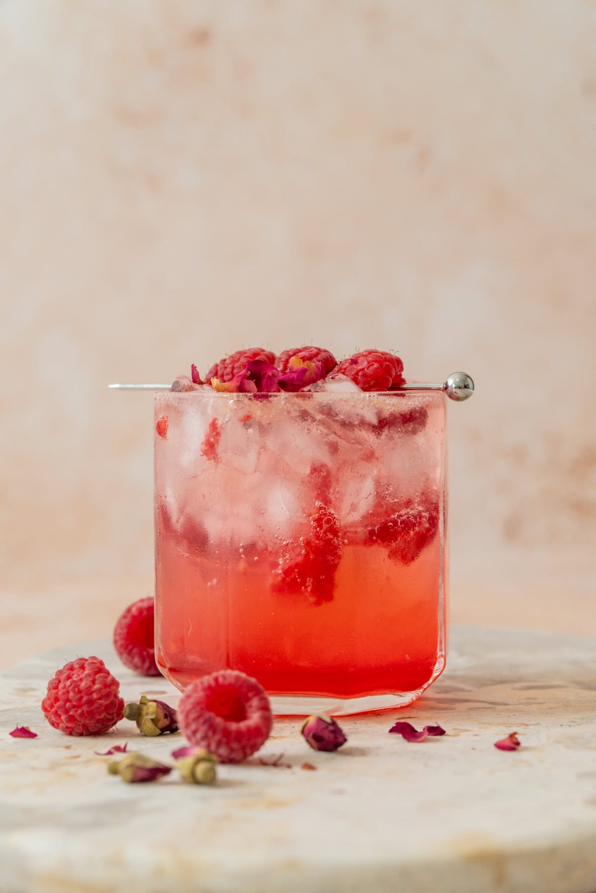 A glass of pink raspberry rose tonic mocktail with ice, raspberries, and rose petals, garnished with a metal pick holding raspberries. Extra raspberries and petals are scattered on a marble surface beside the glass.