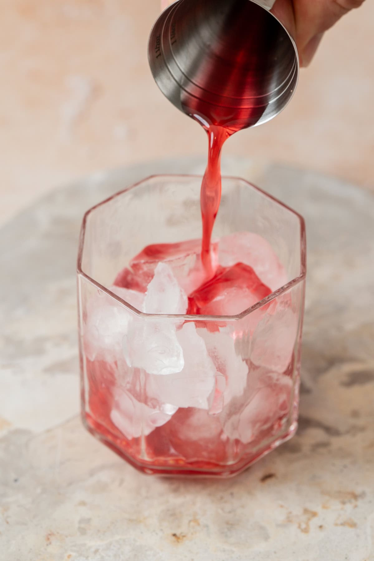 A hand pours a raspberry simple syrup from a metal jigger into a clear glass filled with large ice cubes, set on a light-colored surface.