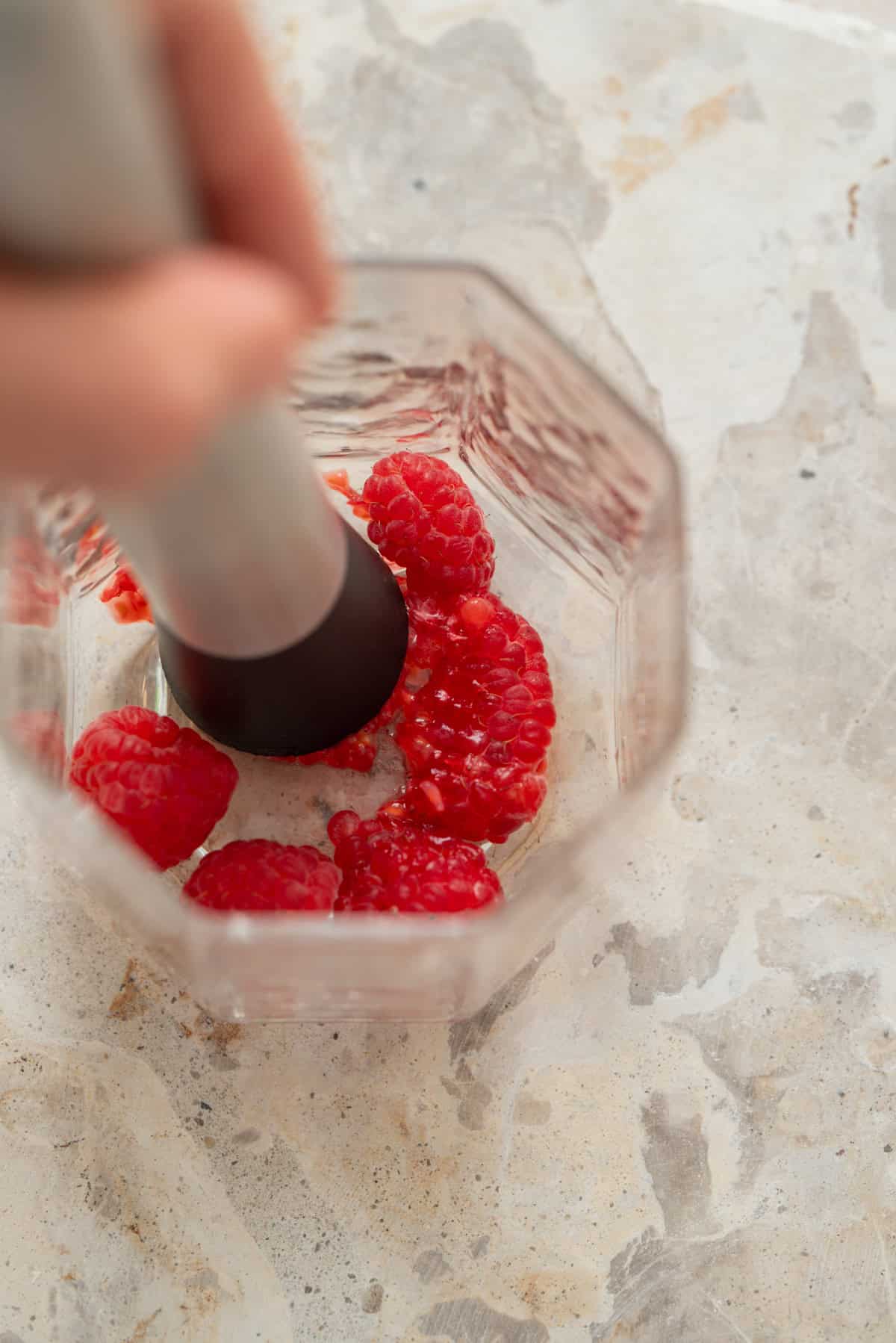 A hand uses a muddler to crush fresh raspberries in a clear glass on a light, textured surface.