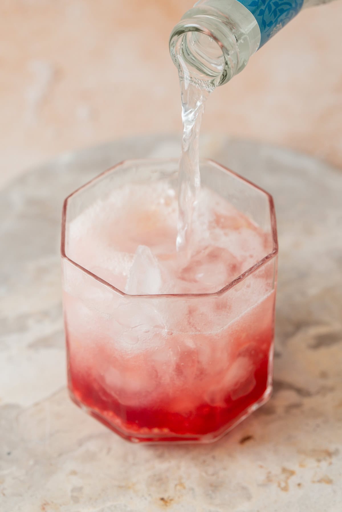 Tonic water is being poured from a bottle into a glass filled with ice and a red liquid on a light-colored surface.
