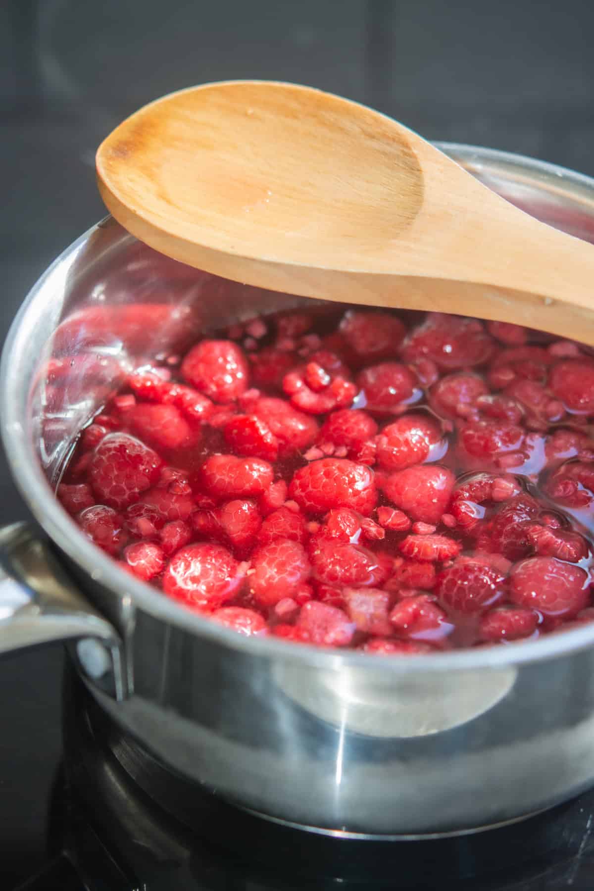 A stainless steel saucepan filled with raspberries and liquid is being heated on a stovetop, with a wooden spoon resting on top of the pan.