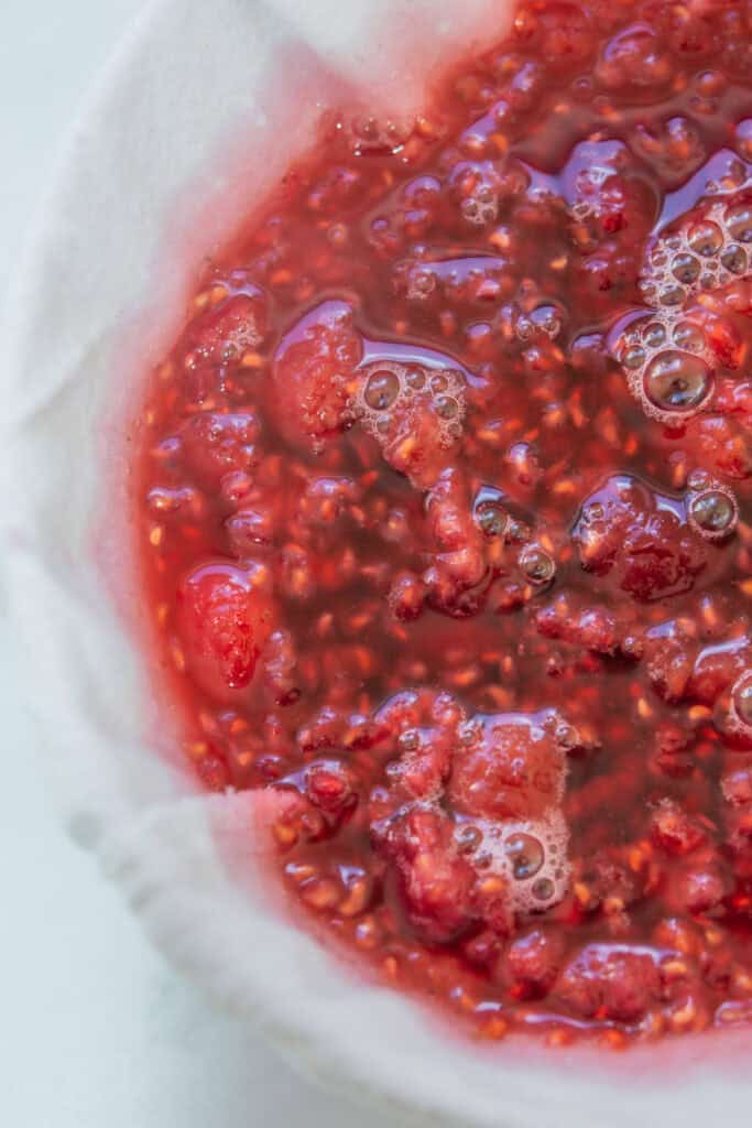 Close-up of mashed raspberries and juice in a white cloth, likely being strained for jam or syrup, with visible seeds and bubbles on the surface.