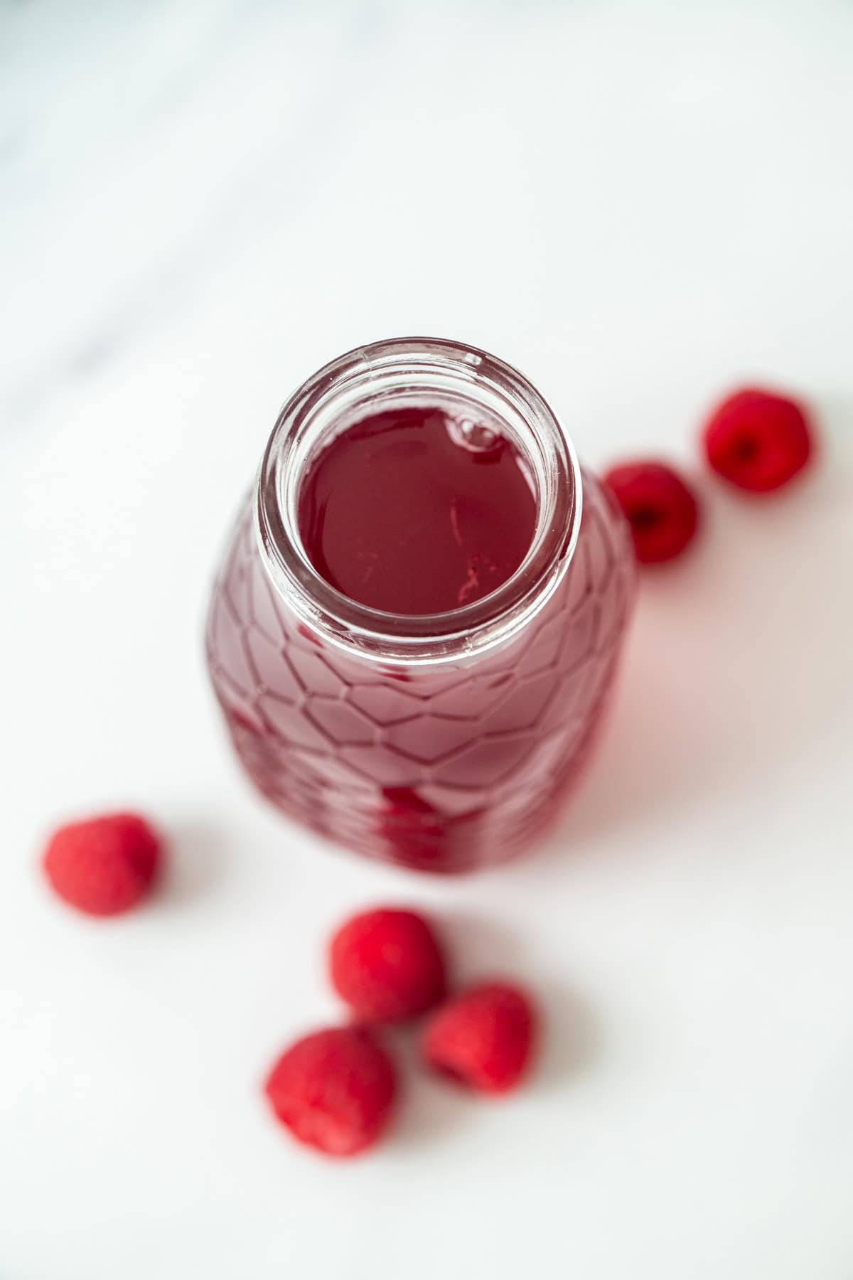 A glass bottle filled with red raspberry simple syrup sits on a white surface, surrounded by several fresh raspberries.