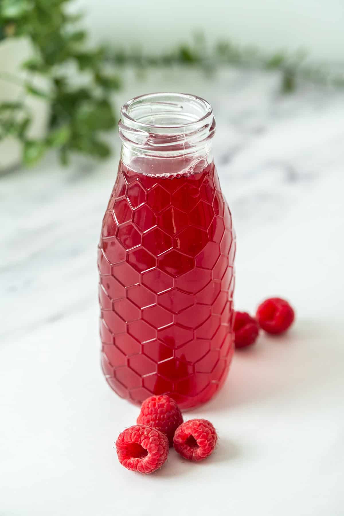 A clear glass bottle with a honeycomb pattern filled with raspberry simple syrup, sits on a white surface next to several fresh raspberries. Green leaves are blurred in the background.
